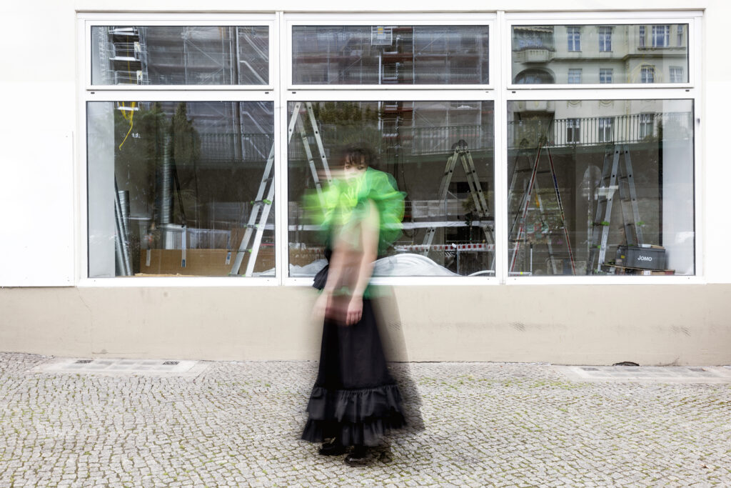 Foto einer Person in dunklem Kleid vorm Fenster eines leeren Ladengeschäfts, draußen, auf der Straße.